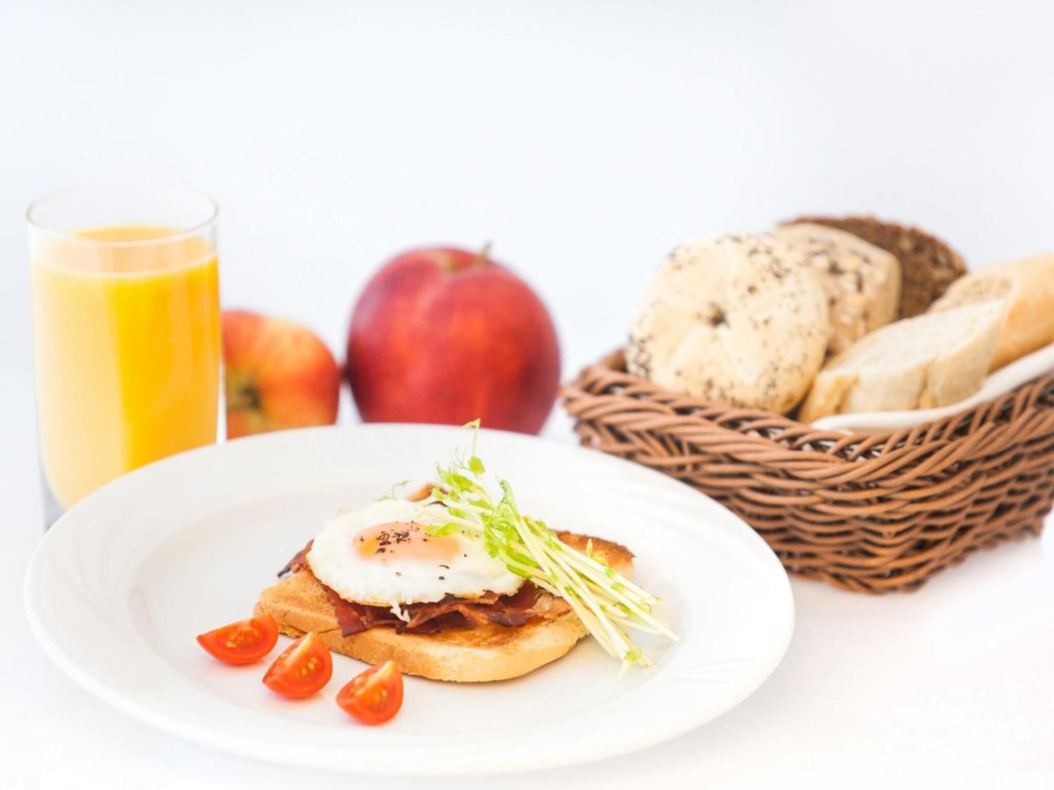 Un plato de desayuno con huevo, tocino y tomate, un vaso de jugo de naranja y una cesta con pan y una manzana.