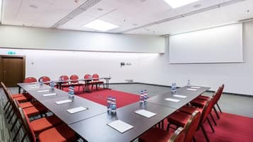 A conference room with a long table, red chairs, water bottles, and a projector screen on the wall.
