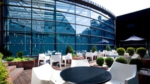 Un espace extérieur aménagé avec des chaises et des tables en plastique blanc, entouré de verdure et bordé d’un bâtiment moderne doté de grandes fenêtres en verre.