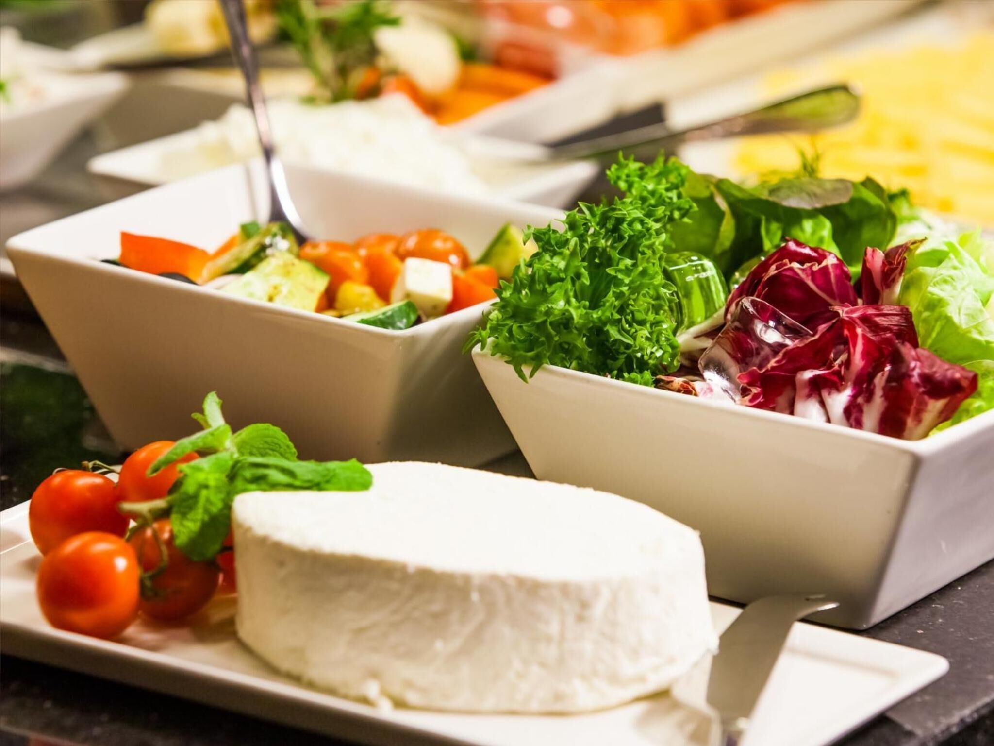 A buffet with various dishes including a bowl of salad, a plate of cheese, and a bowl of mixed vegetables.