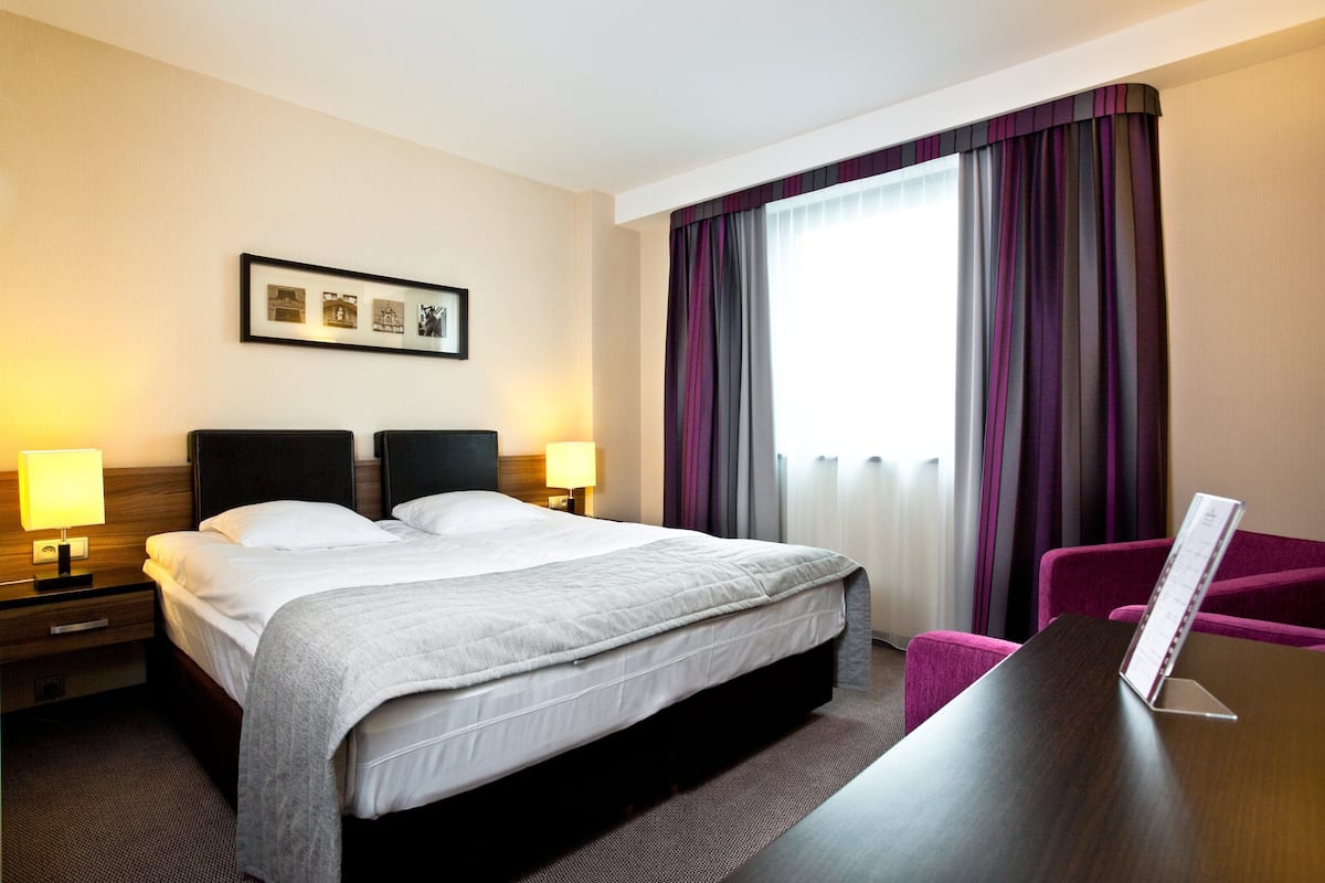 A hotel room with a large bed, two bedside tables with lamps, a framed picture on the wall, a window with purple curtains, and a small table with a menu stand.