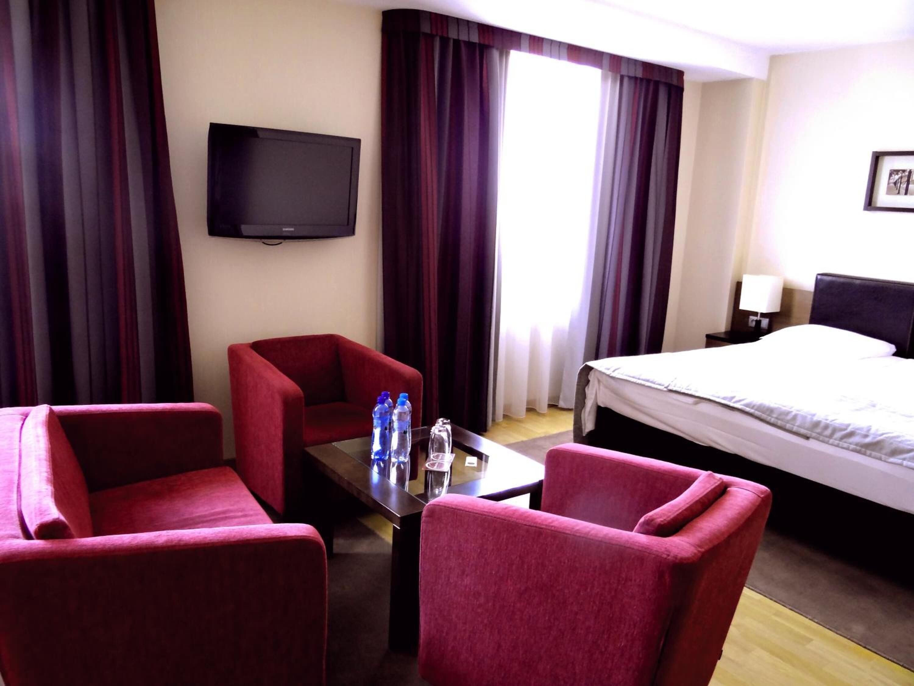 A hotel room with a bed, two red armchairs, a small table with water bottles, a TV on the wall, and a window with curtains.