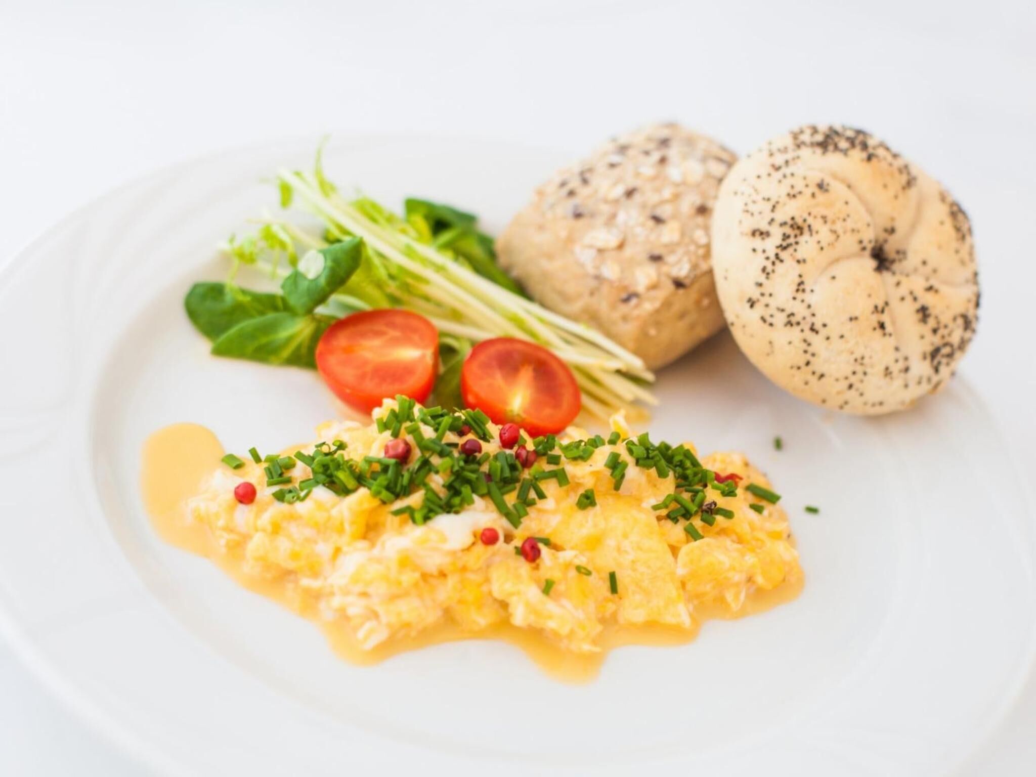 A plate with scrambled eggs, cherry tomatoes, green onions, and a bagel with poppy seeds.