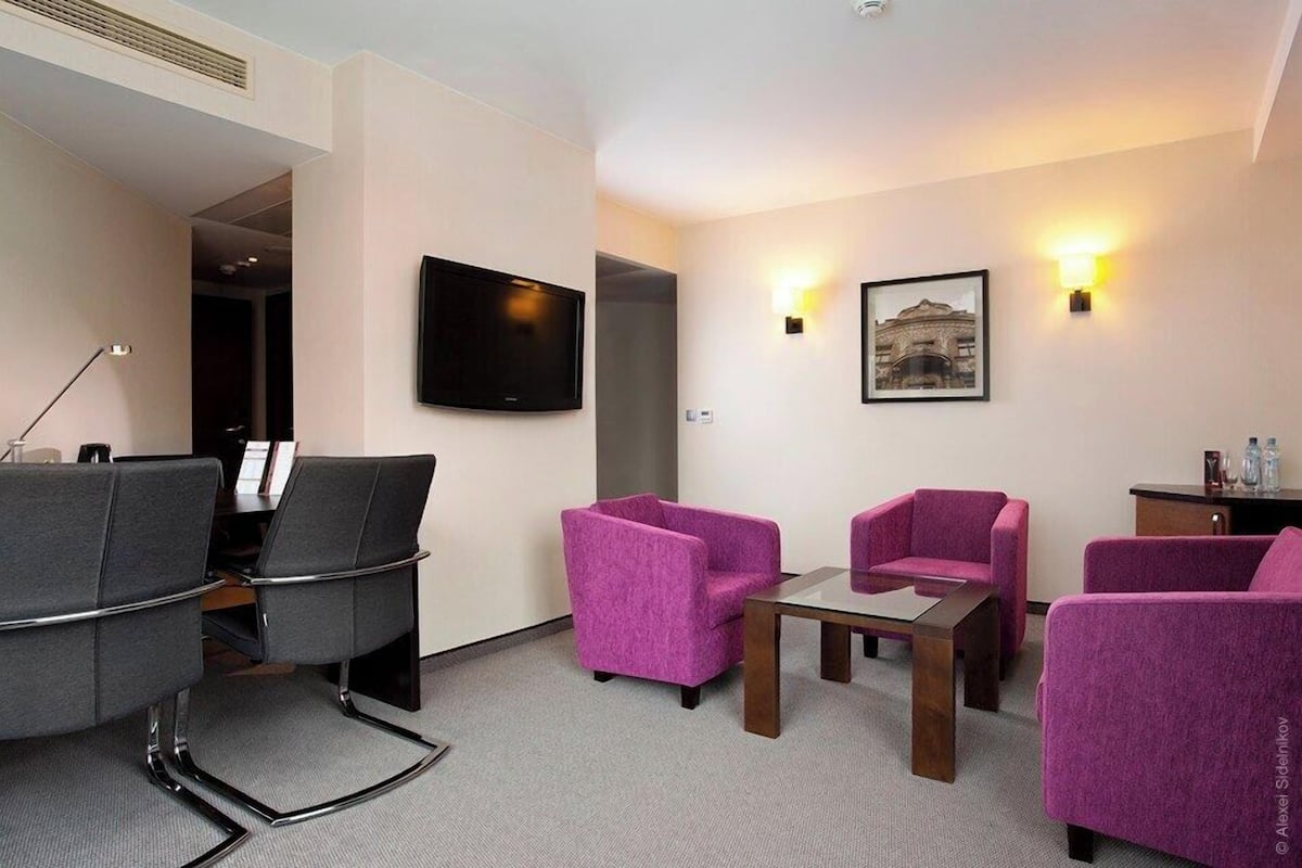 A hotel room with a seating area featuring purple chairs, a flat-screen TV mounted on the wall, and a framed picture above the TV.