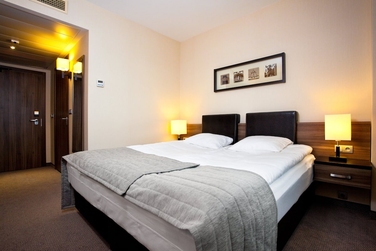 A hotel room with a large bed, two bedside tables with lamps, a framed picture on the wall, and a wooden door.