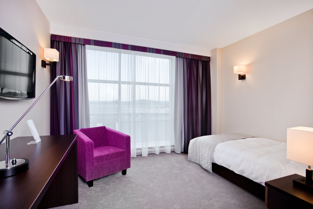 A hotel room with a bed, a desk with a lamp, a chair, and a TV mounted on the wall.