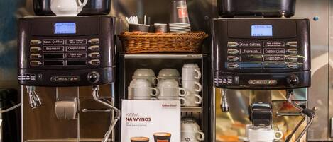 Two coffee machines with options for Kawa Coffee, Espresso, and Cappuccino. There are cups and a sign for Quous Hotel.
