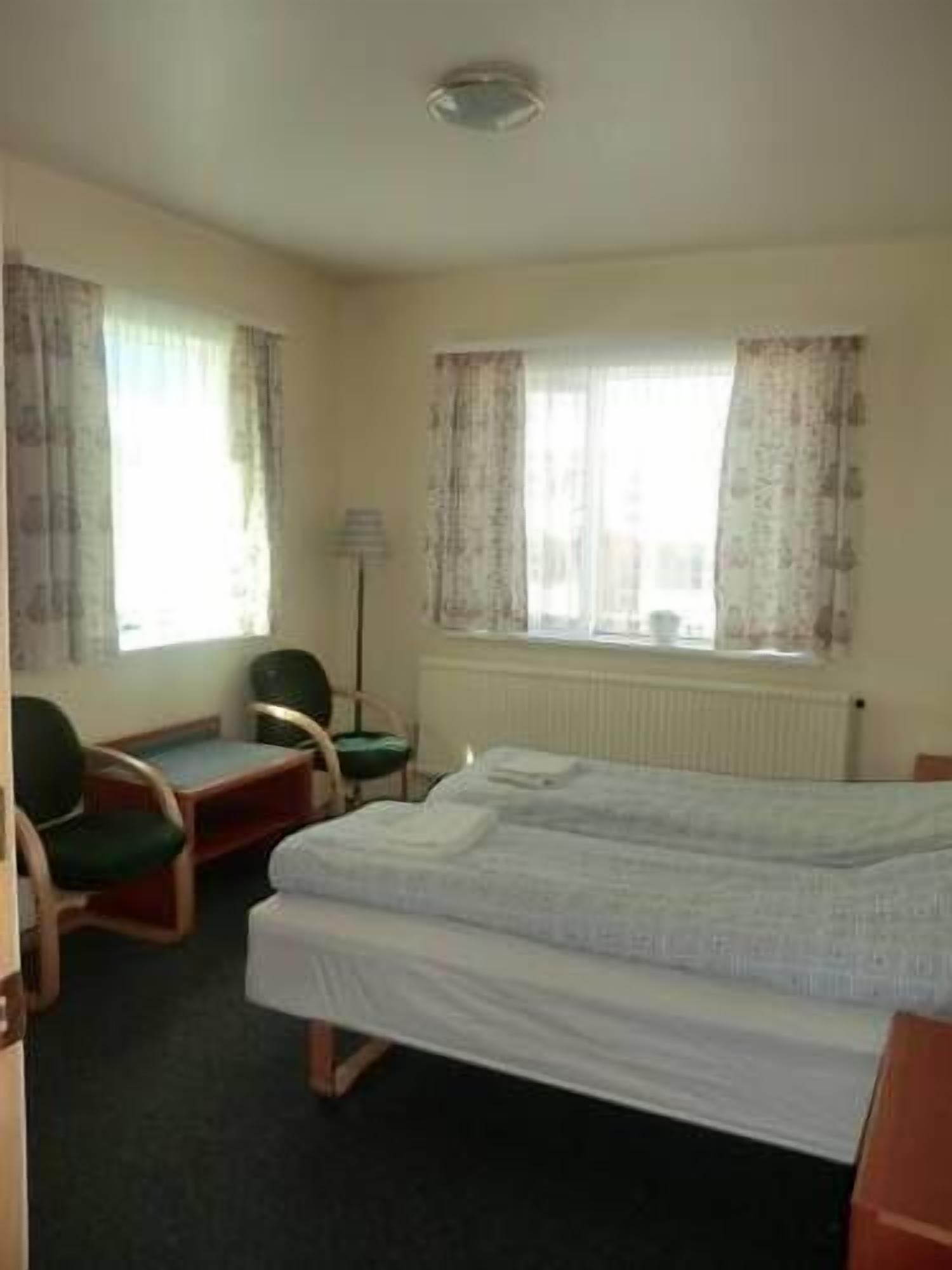 Une chambre d’hôtel avec un lit simple, deux chaises, une petite table et une fenêtre avec des rideaux.