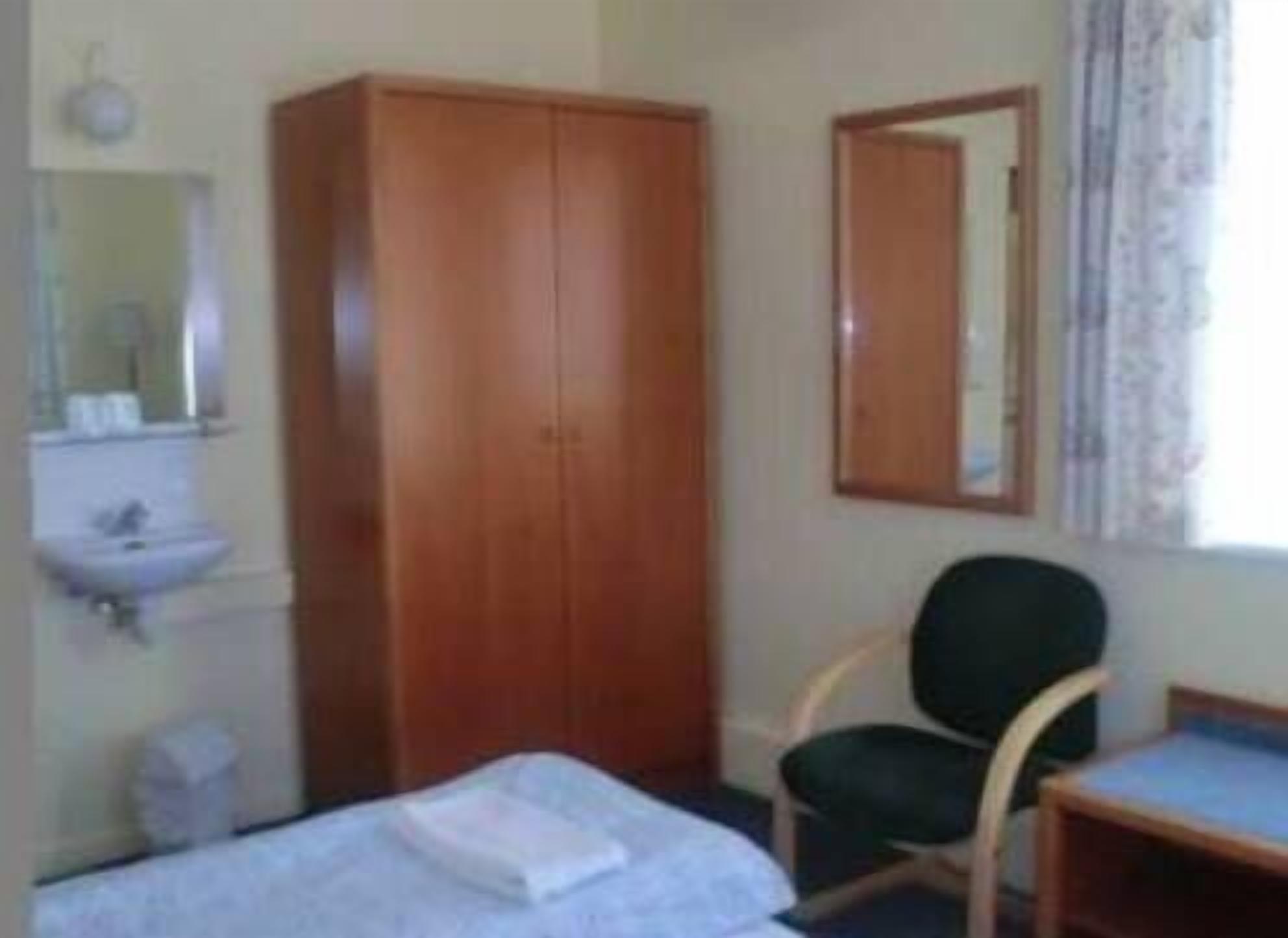 Une petite pièce simple, avec une armoire en bois, un lit simple, une chaise, un bureau et un miroir.