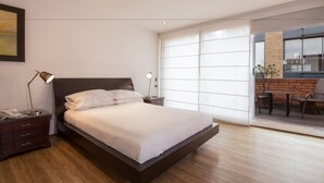 Une chambre moderne avec un grand lit, des tables de chevet, un bureau et une vue sur un espace extérieur agrémenté d’un mur en briques et de chaises.
