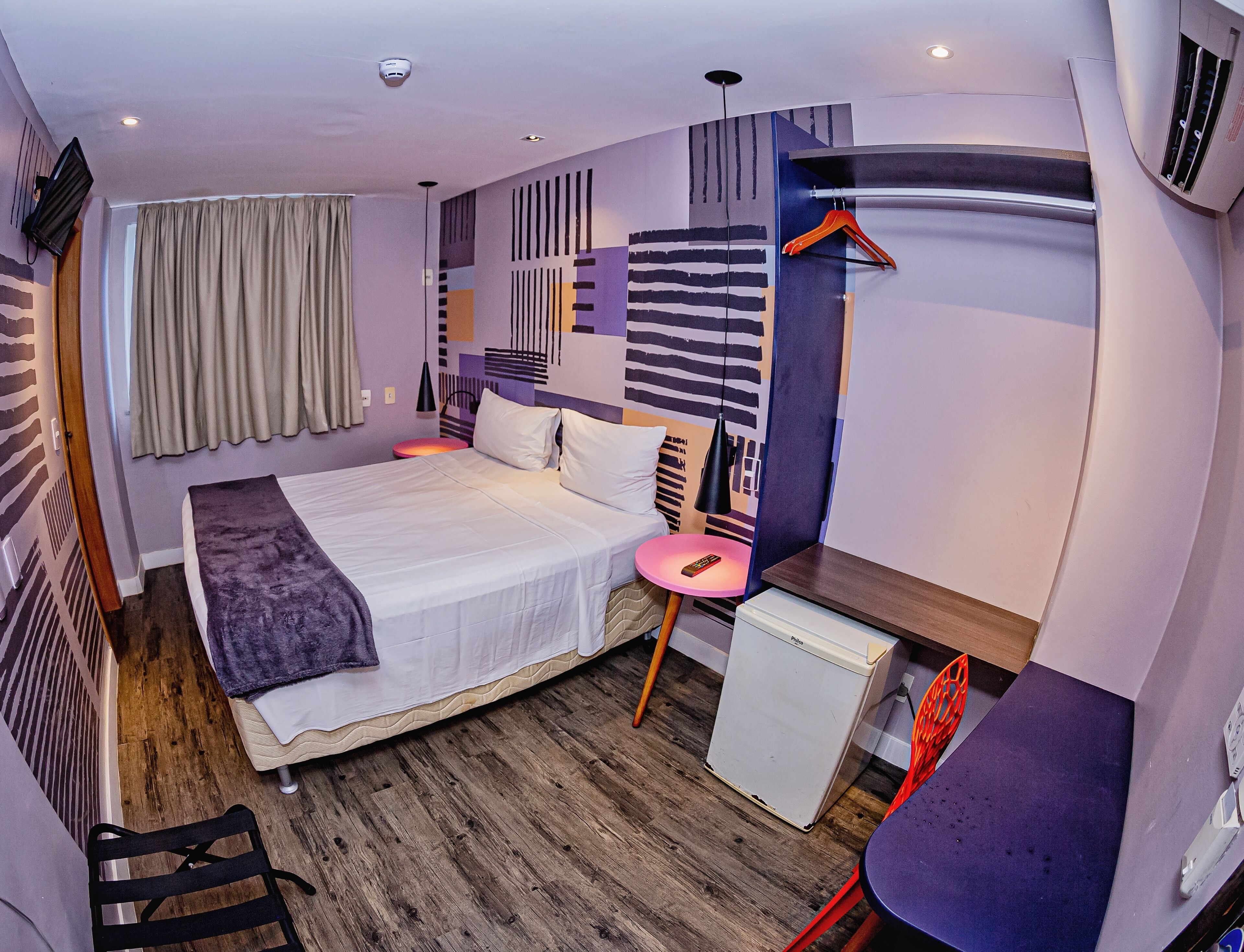 A modern hotel room with a large bed, a desk, a mini fridge, and a hanging coat rack.