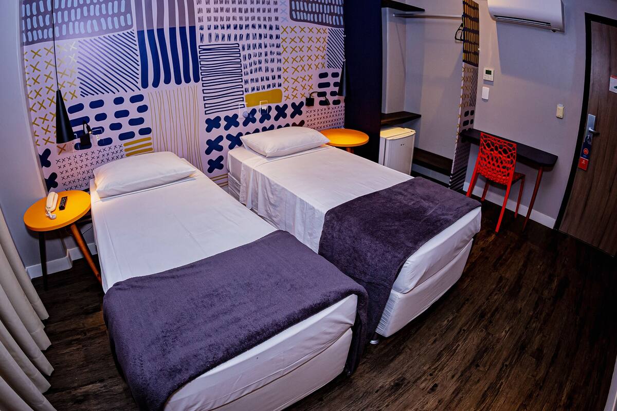 A hotel room with two beds, a small table with a lamp, a red chair, and a wall air conditioner.