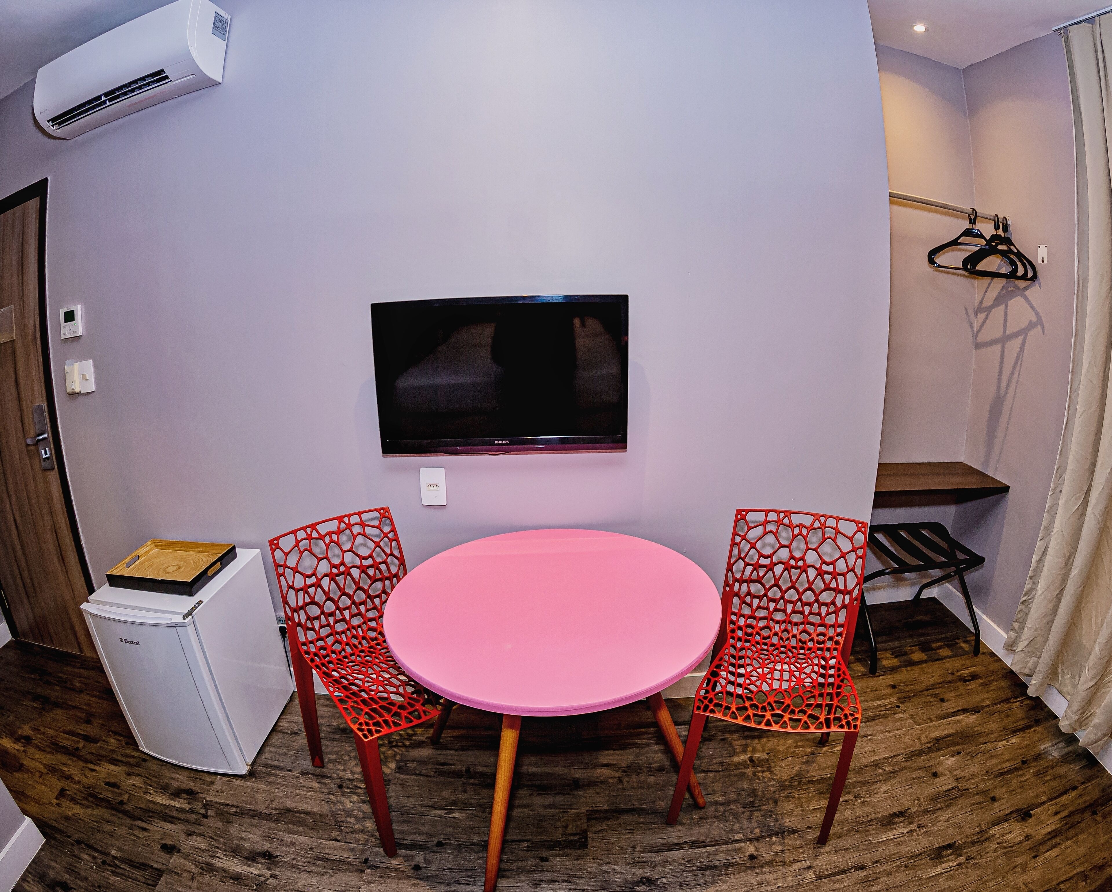 Una habitación con una mesa rosa, dos sillas rojas, un televisor de pantalla plana, un refrigerador blanco pequeño y suelo de madera.