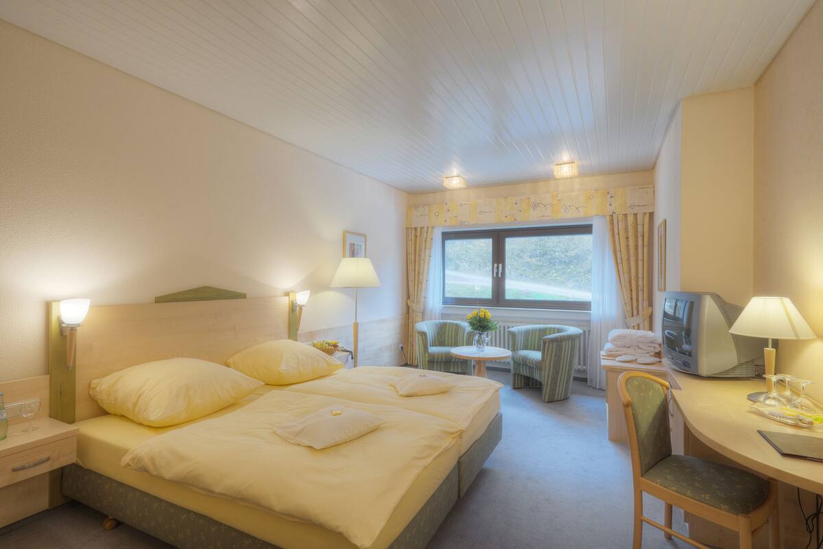 Una habitación de hotel con cama, lámparas de noche, un escritorio con computadora, una silla y una ventana con cortinas.