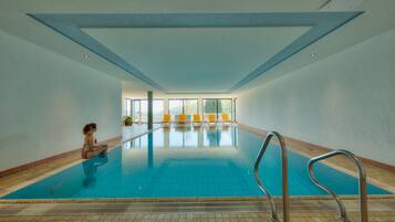 An indoor swimming pool with a clear view of the pool area, a person sitting by the pool, and a row of yellow chairs in the background.
