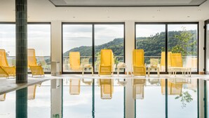 An indoor swimming pool with yellow chairs and large windows offering a view of a forested hill. - Waldhotel Sonnenberg (Bollendorf)