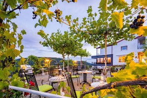 An outdoor seating area with tables and chairs, surrounded by grapevines and a building in the background. - Waldhotel Sonnenberg (Bollendorf)