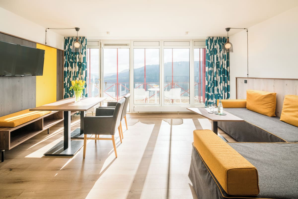 Una sala de estar moderna con un sofá, una mesa de comedor y sillas. Hay un televisor montado en la pared, y la habitación tiene ventanas grandes con cortinas.