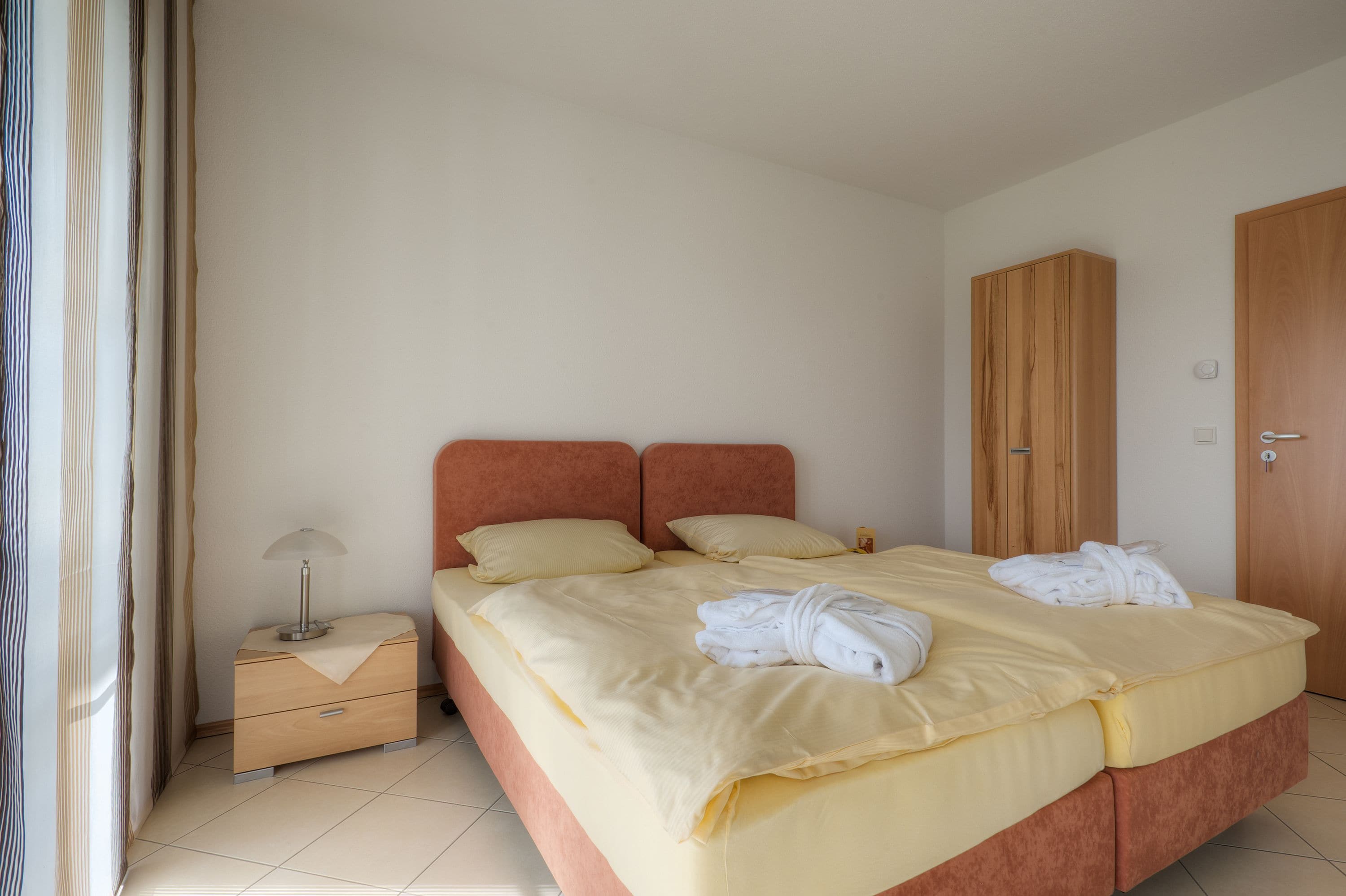 Una cama bien hecha con colcha y almohadas amarillas, una mesita de noche de madera con una lámpara, y un armario de madera.