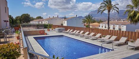 A rooftop pool with lounge chairs, a wooden deck, and a view of buildings and mountains.