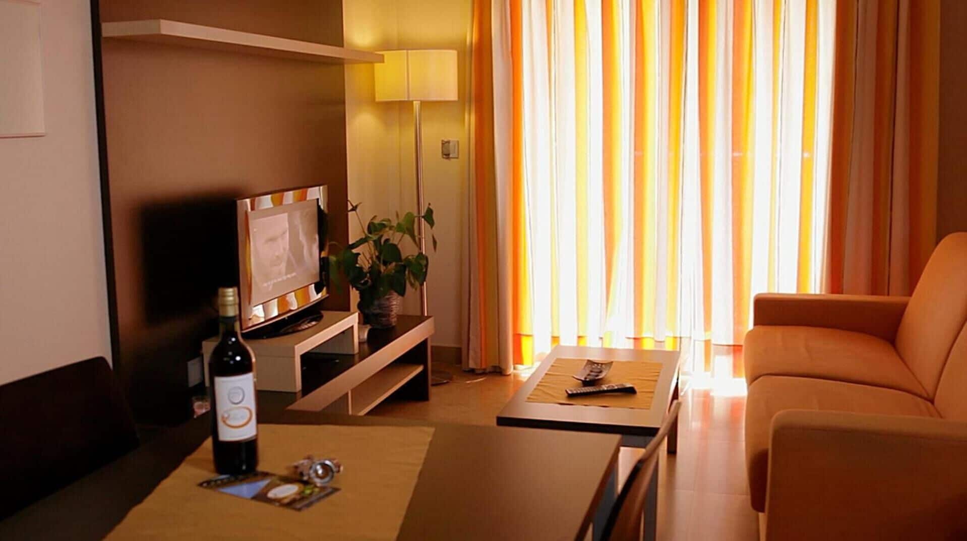 A hotel room with a TV, a bottle of wine, a plant, a sofa, and a table.
