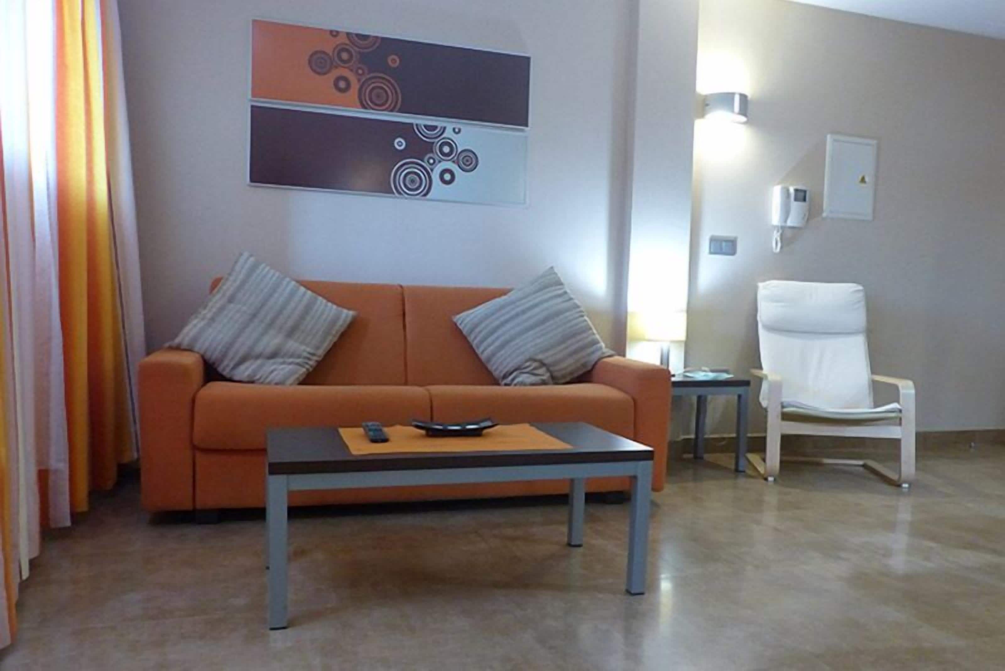 Ett modernt vardagsrum med en orange soffa, två kuddar, ett soffbord med en fjärrkontroll, en vit fåtölj och en väggmonterad lampa.