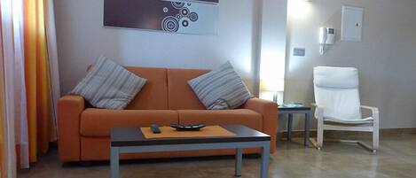 Ett modernt vardagsrum med en orange soffa, två kuddar, ett soffbord med en fjärrkontroll, en vit fåtölj och en väggmonterad lampa.