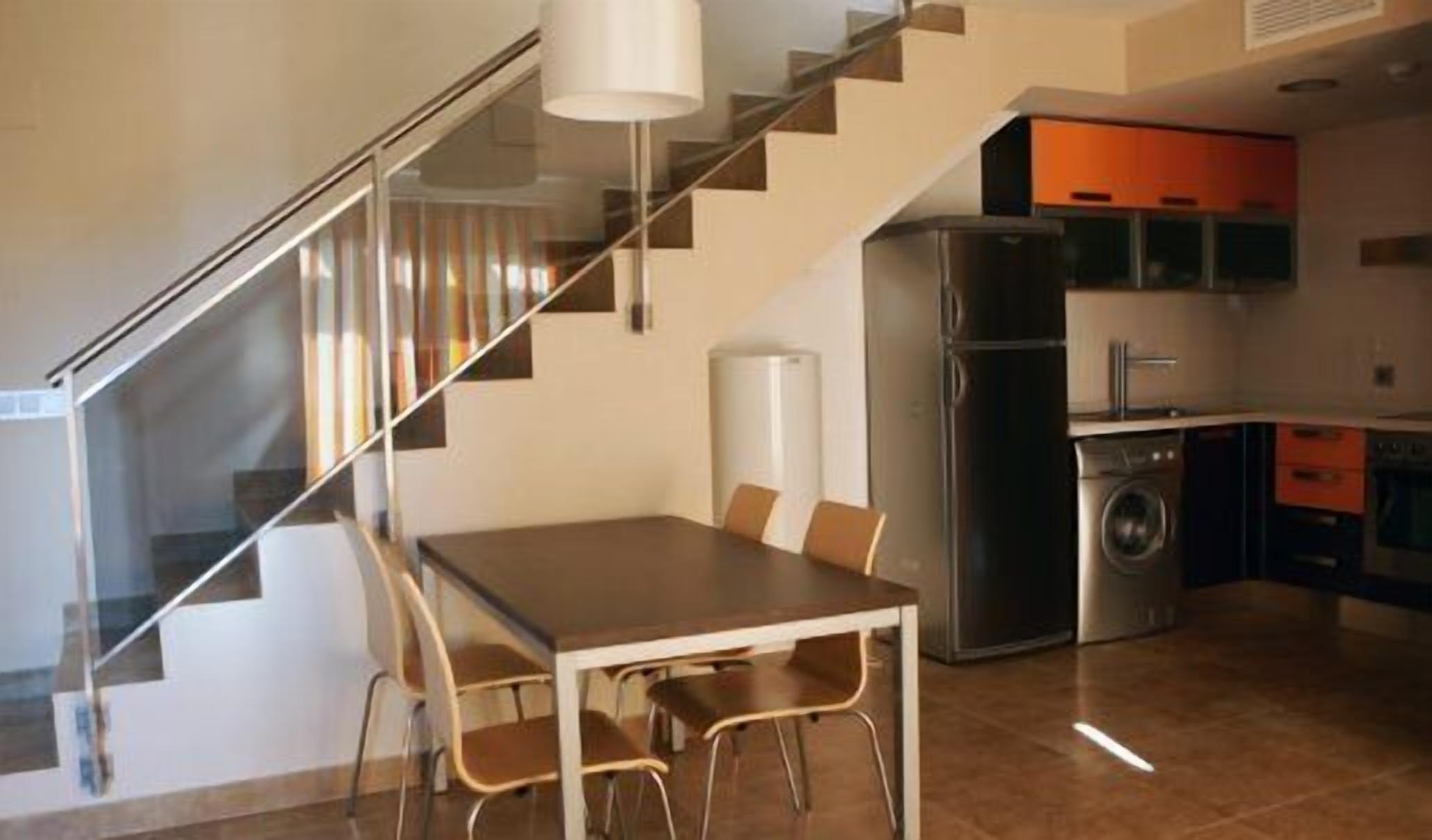 A modern kitchen with stainless steel appliances, a dining area with a table and chairs, and a staircase with glass railings.