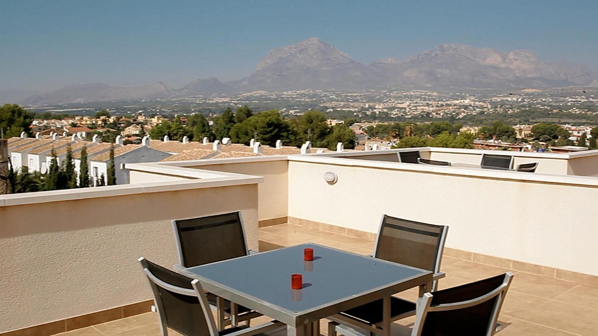 A rooftop terrace with a table and chairs, a view of a mountain range, and a cityscape in the background.