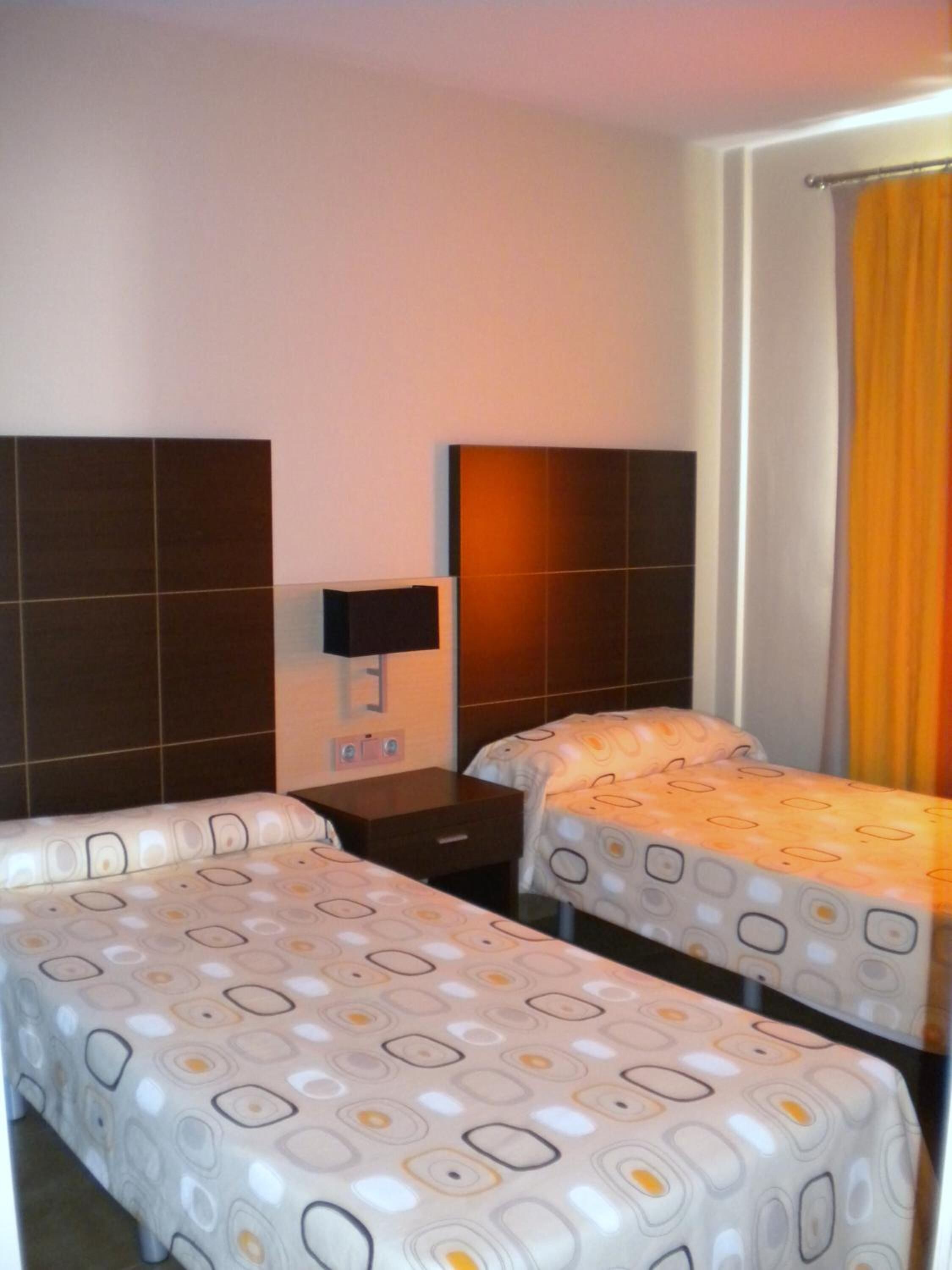 Habitación de hotel con dos camas, una mesita de noche, un televisor montado en la pared y una ventana con cortinas.