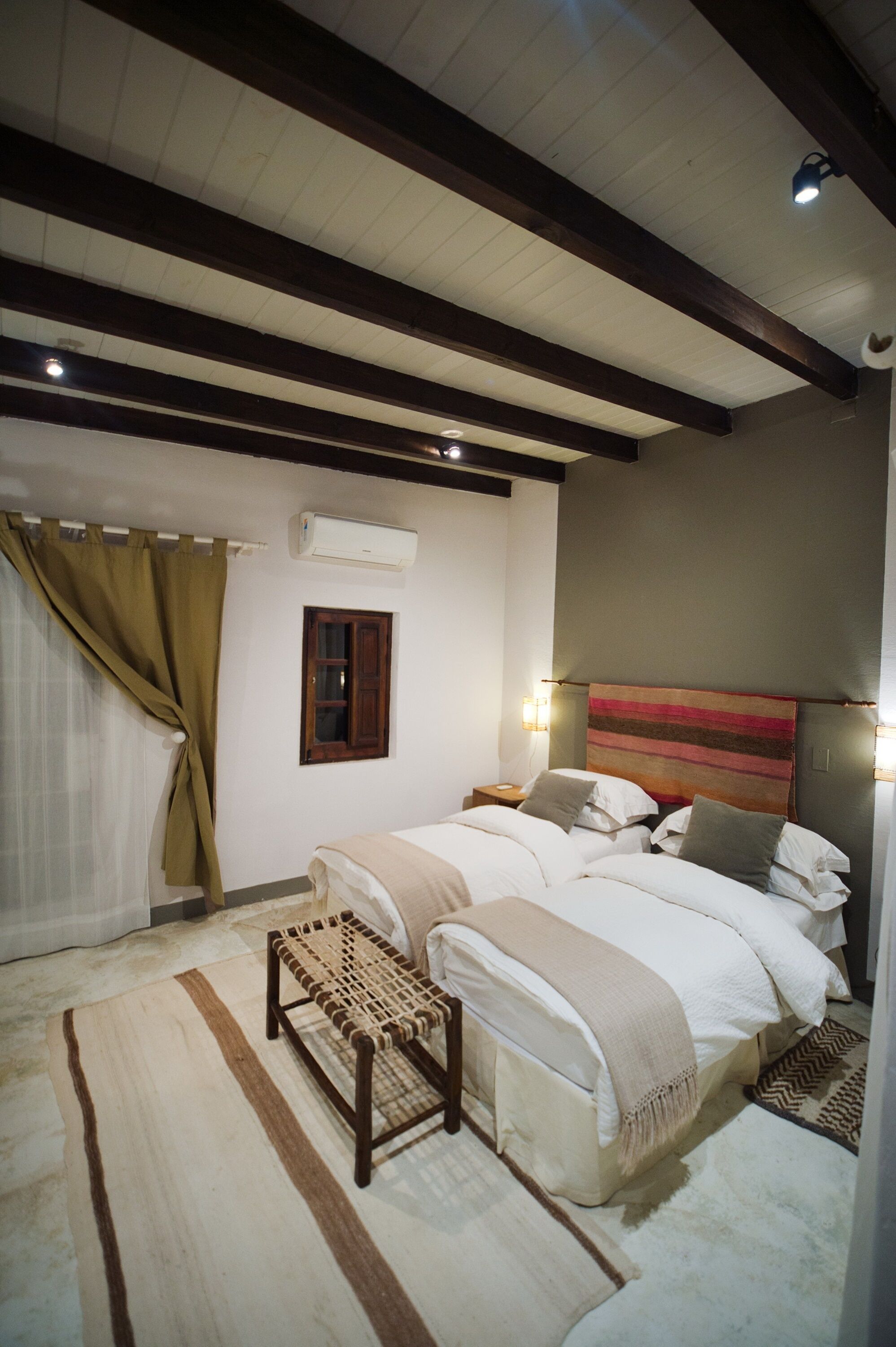 A bedroom with a bed, bedside tables, a bench, a window, and a wall-mounted air conditioner.