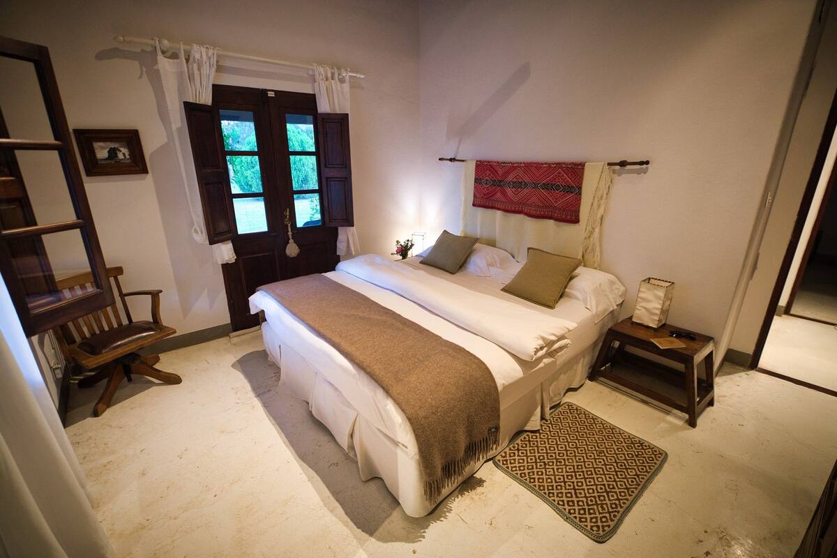 A bedroom with a bed, a wooden chair, a nightstand, a patterned rug, and a window with curtains.
