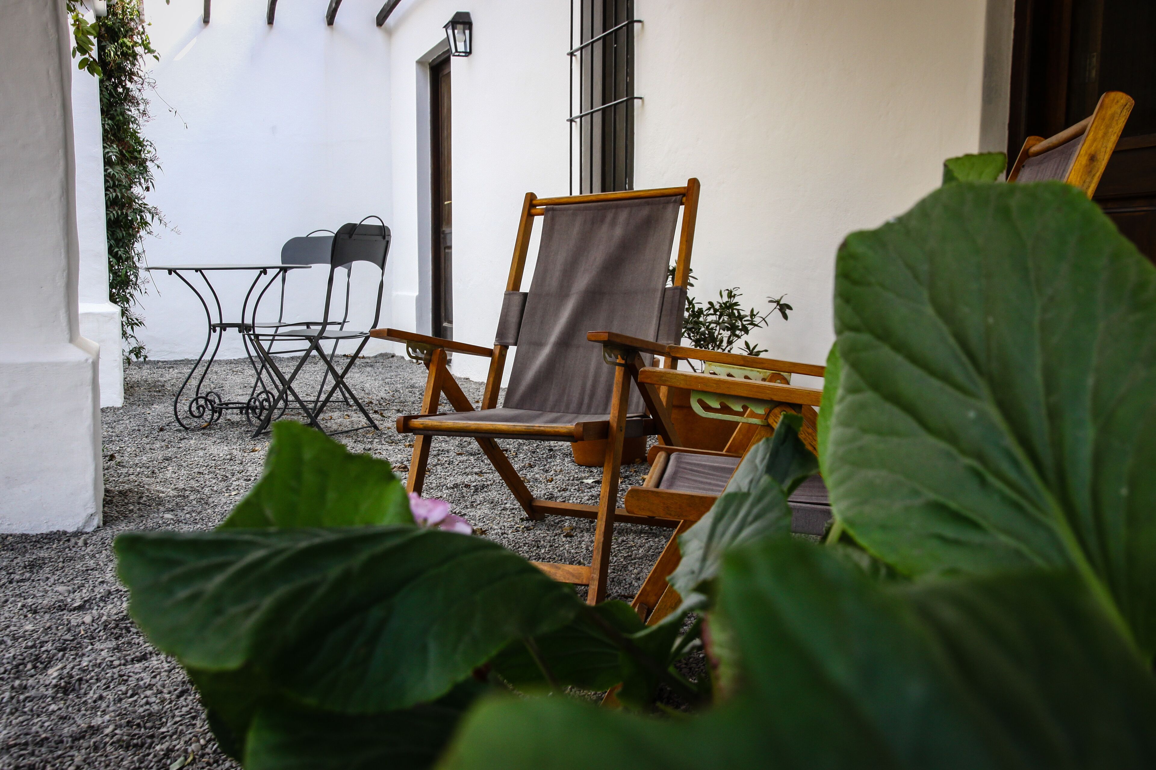 Een terras met een houten stoel, een kleine tafel en een metalen stoel. In de voorgrond staat een plant en op de achtergrond een witte muur.