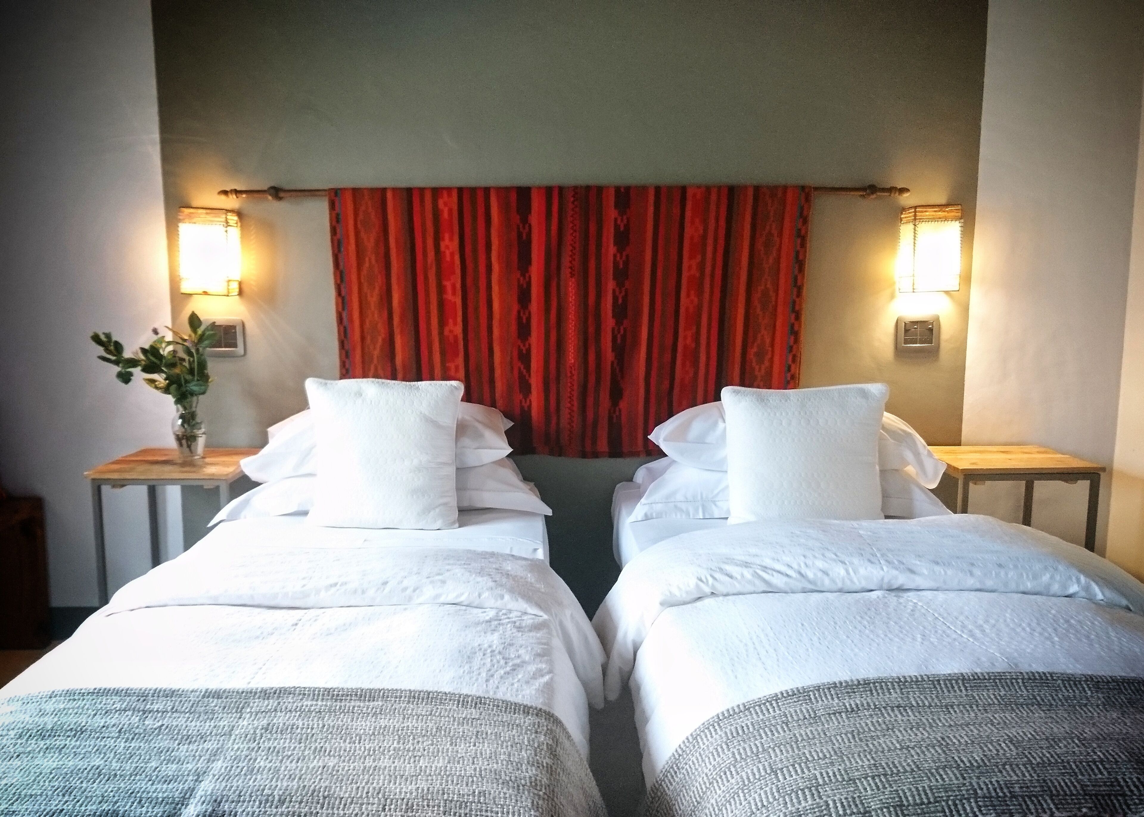 Habitación doble con dos camas, cada una con ropa de cama y almohadas blancas. Hay dos mesitas de noche, una a cada lado de la cama. Un jarrón con flores está sobre la mesita de la izquierda. La habitación tiene una cortina roja y dos lámparas de pared a cada lado de la cortina.