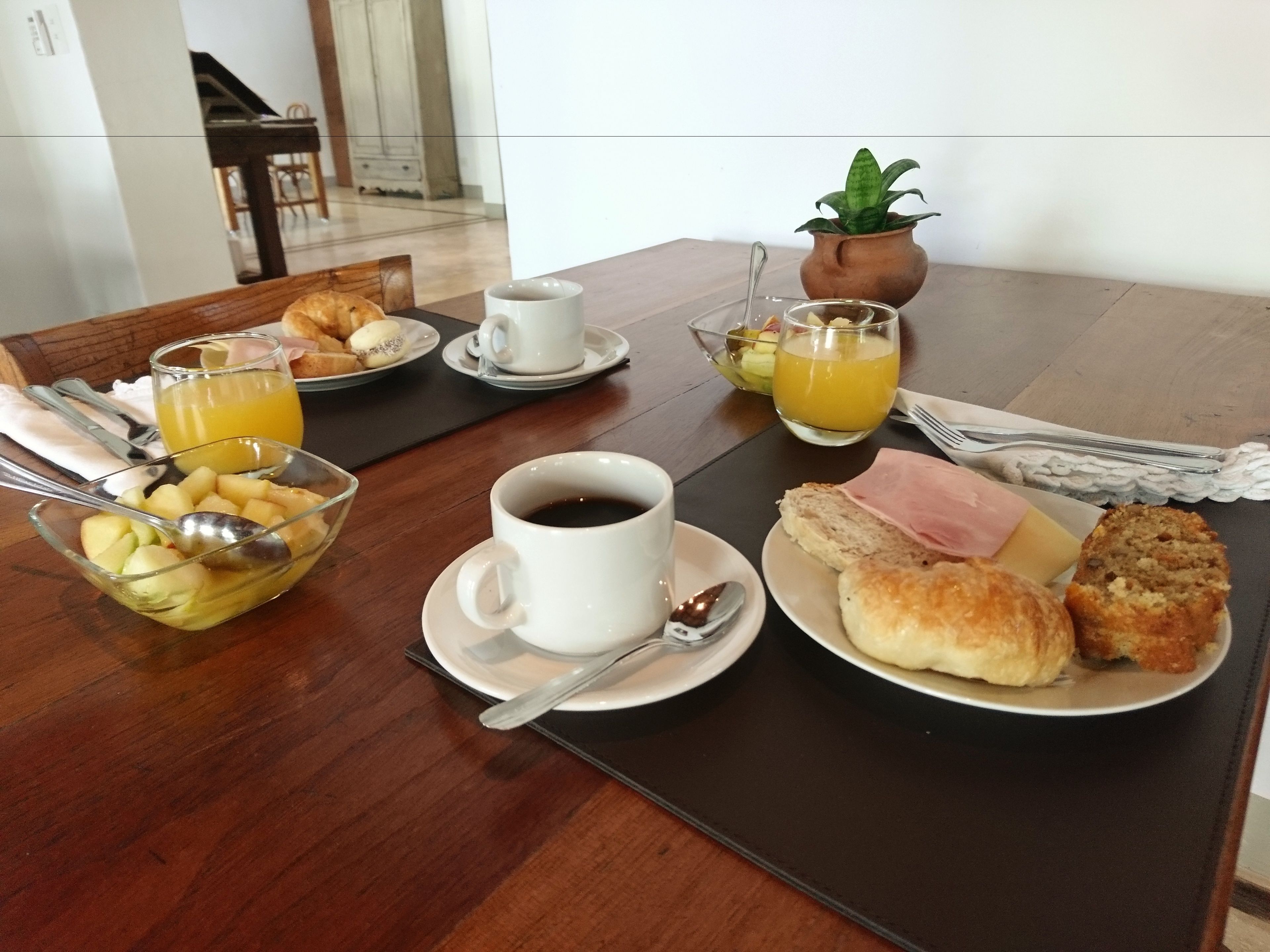 Uma mesa de café da manhã com uma tigela de frutas, uma xícara de café, um copo de suco de laranja e um prato com doces.