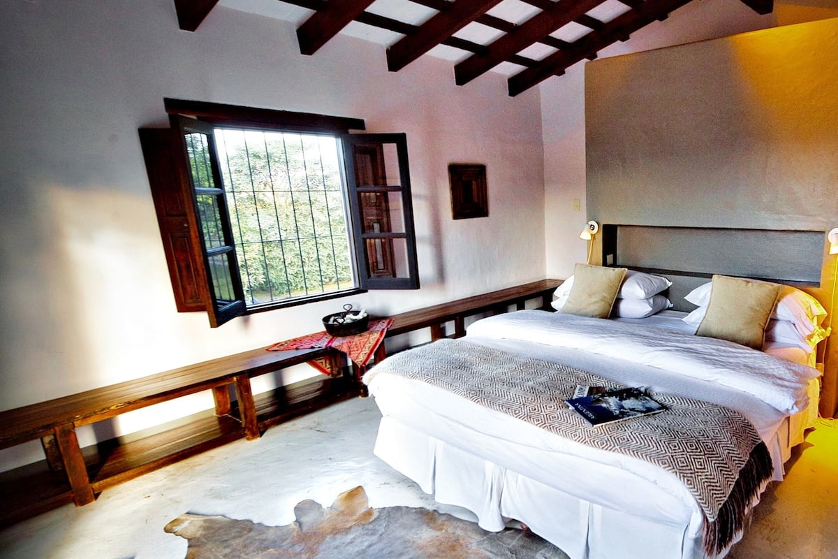 A bedroom with a large bed, a bench, a rug, and a window with a view of greenery.