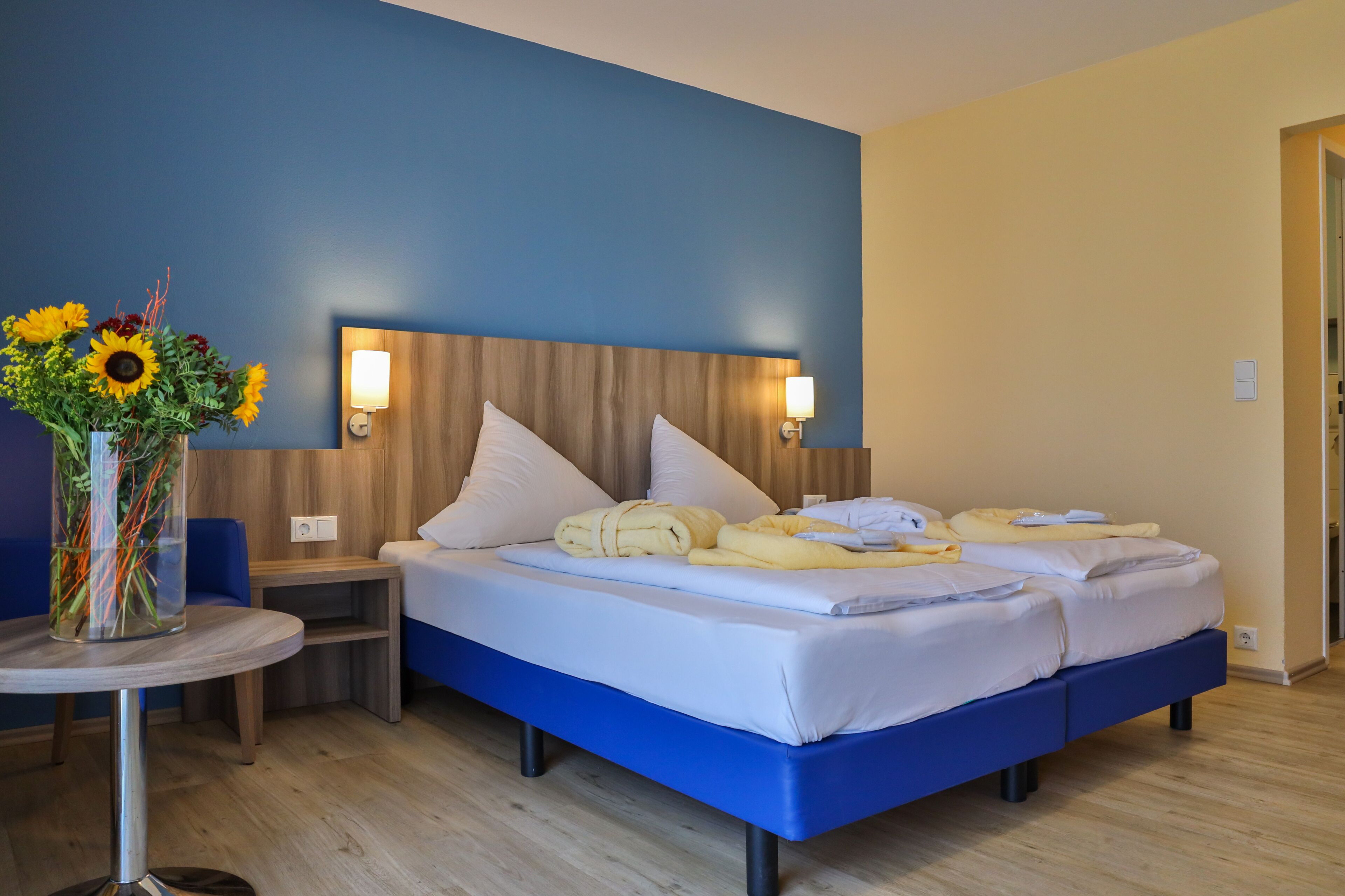A hotel room with two beds, a wooden headboard, bedside lamps, a small table with a vase of flowers, and a blue accent wall.