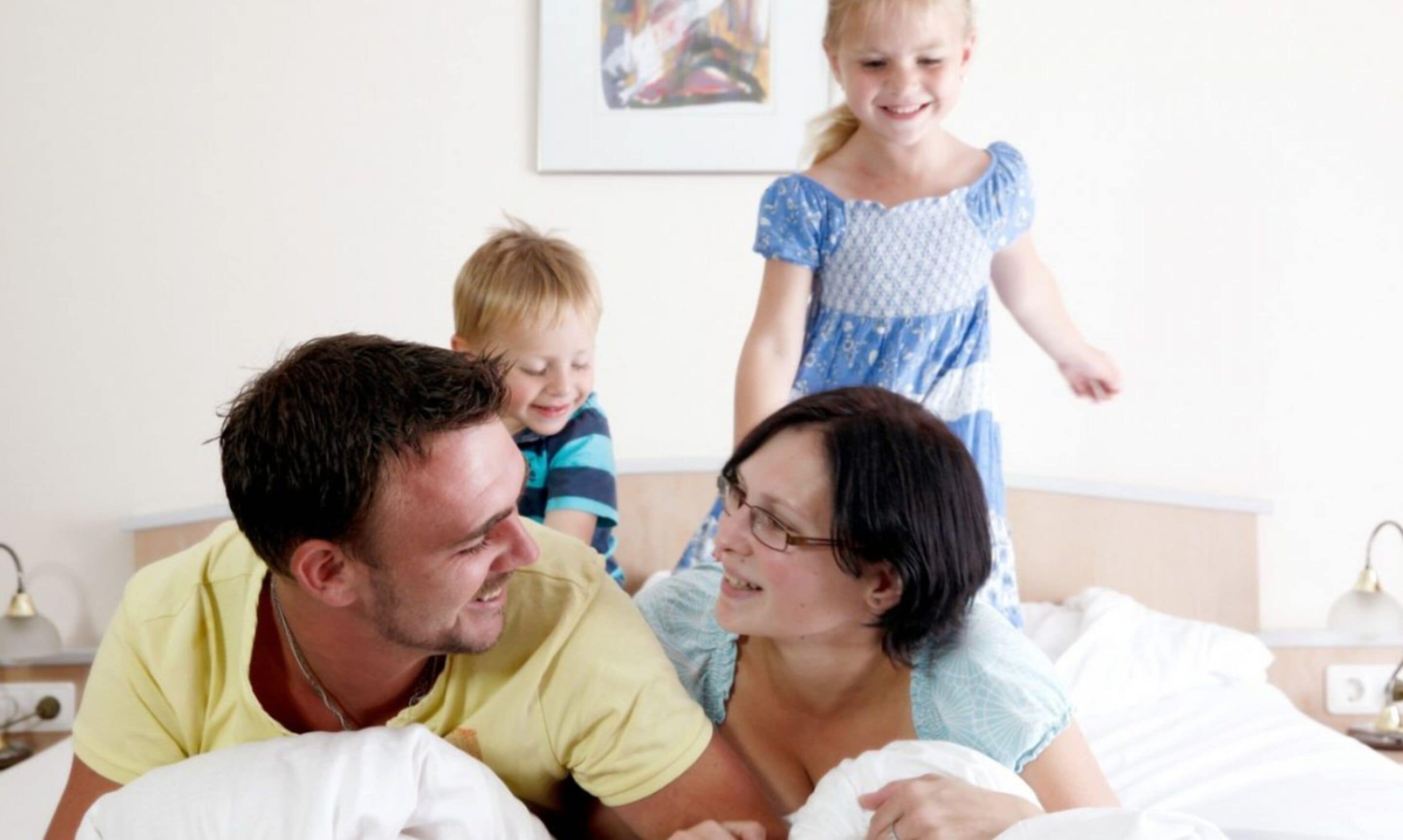 A family in a bedroom with a bed, bedside table, lamp, and a painting on the wall.