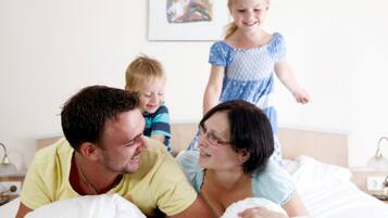 A family in a bedroom with a bed, bedside table, lamp, and a painting on the wall.