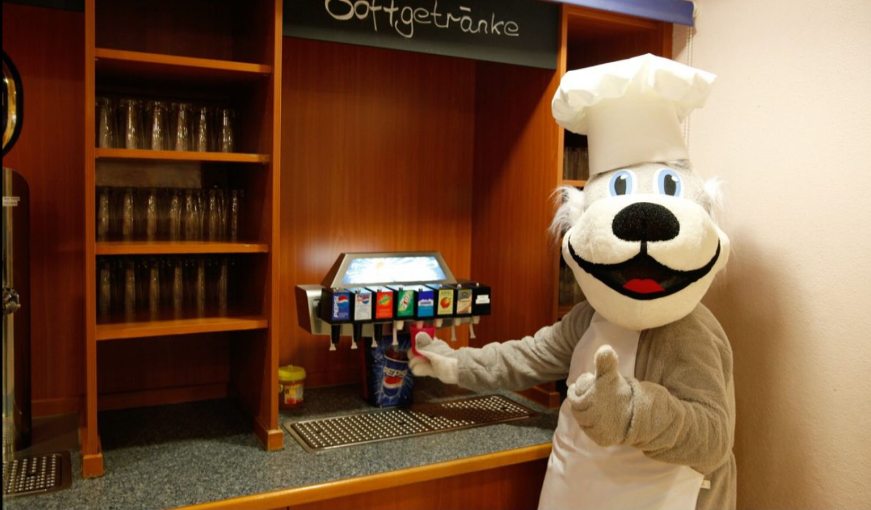 Un personaje promocional vestido de cocinero frente a un dispensador de bebidas con diversas opciones.