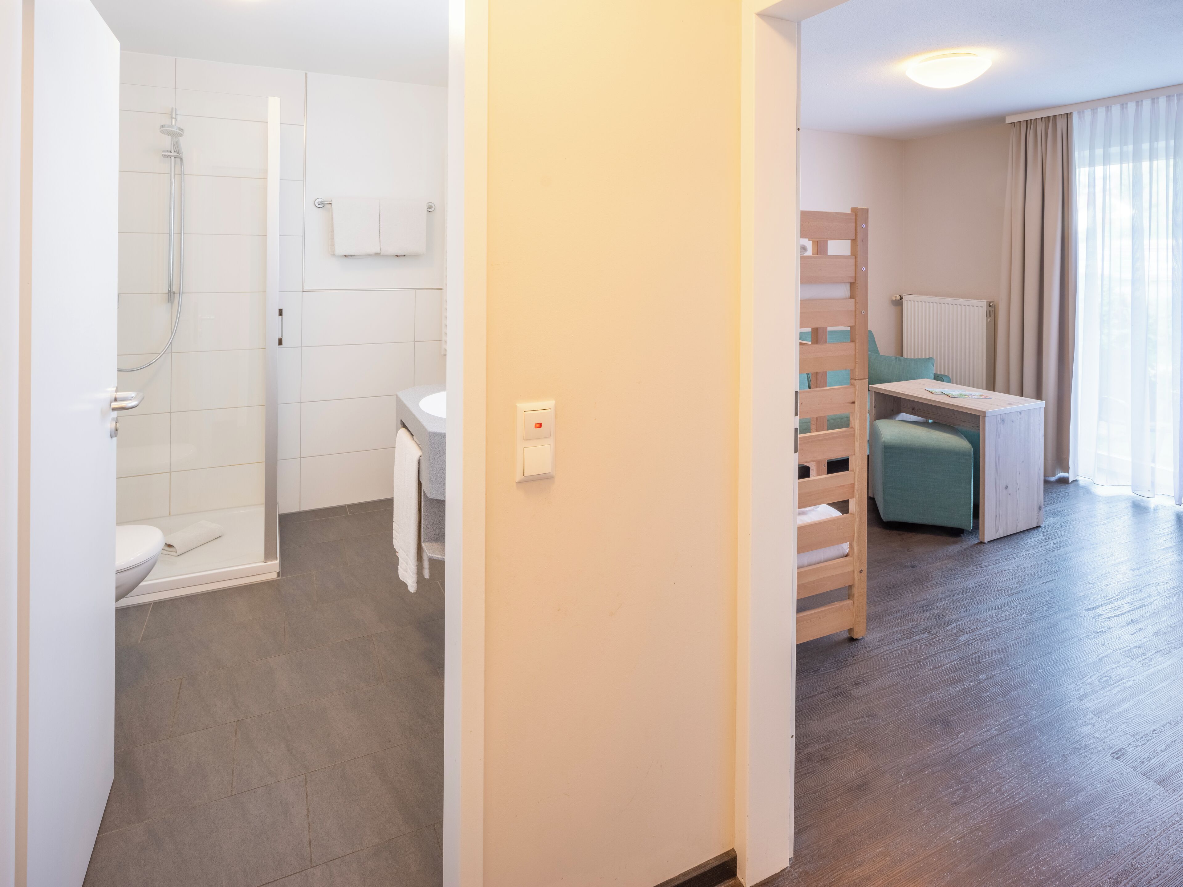 A modern bathroom with a shower, toilet, and sink. There is a wooden bed frame and a small desk in the room.
