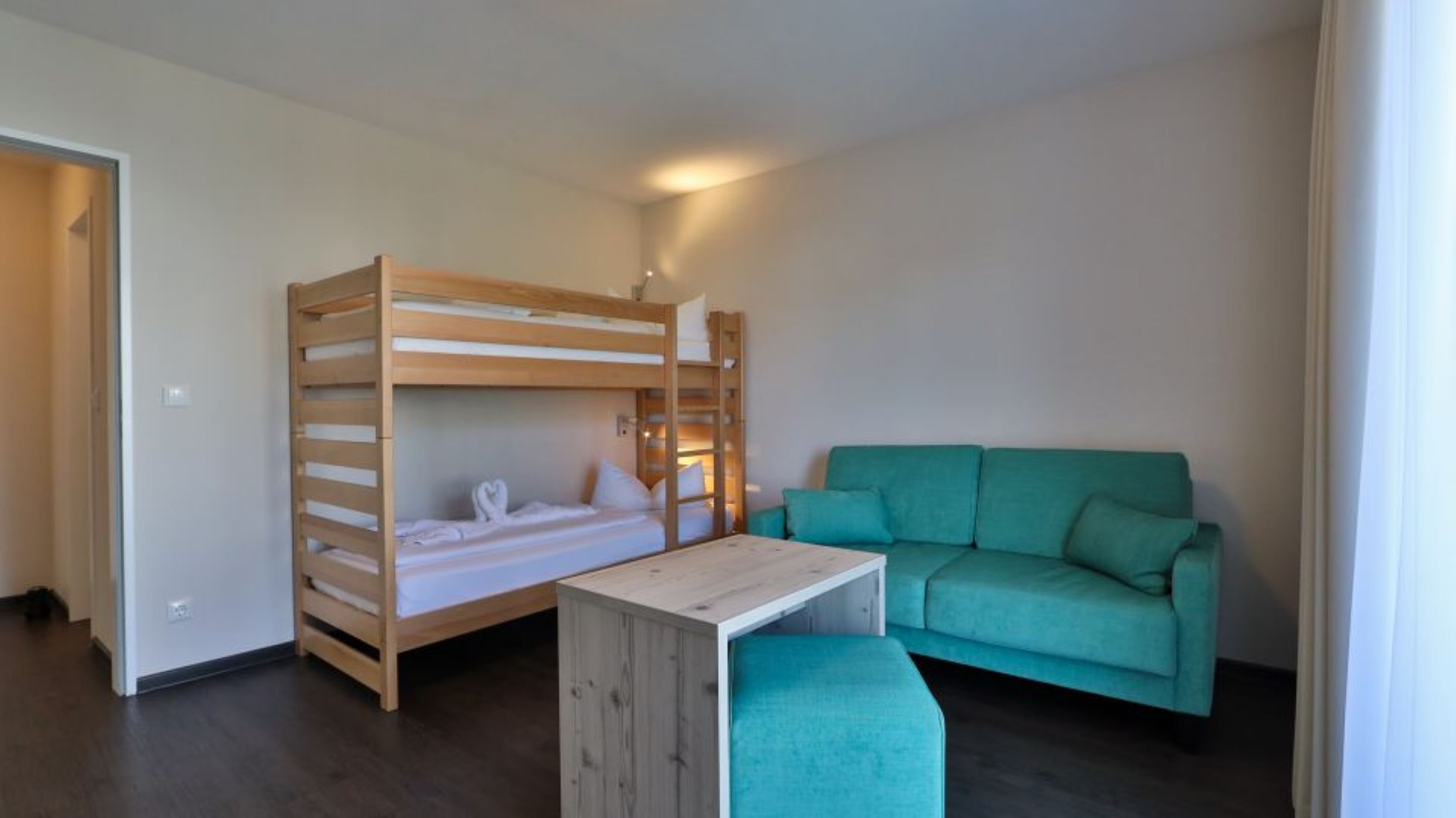 Een kleine, goed verlichte kamer met een stapelbed, een blauwgroene bank en een klein tafeltje.