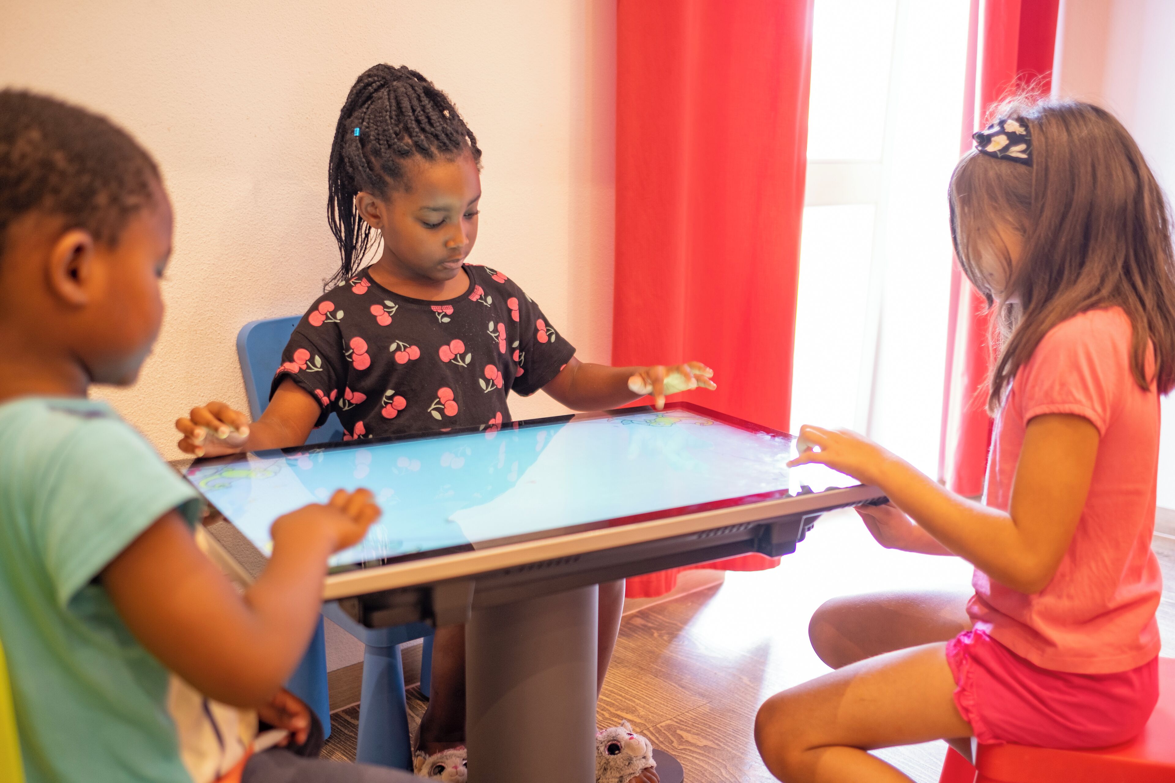 Trois enfants jouent à un jeu sur une tablette, assis autour d’une table.