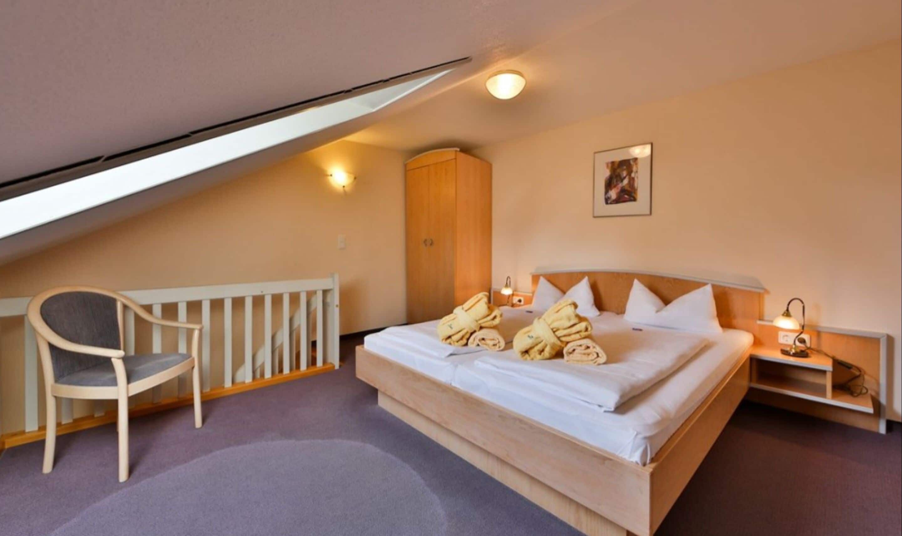 A bedroom with a bed, a chair, a wardrobe, and a small table with a lamp.