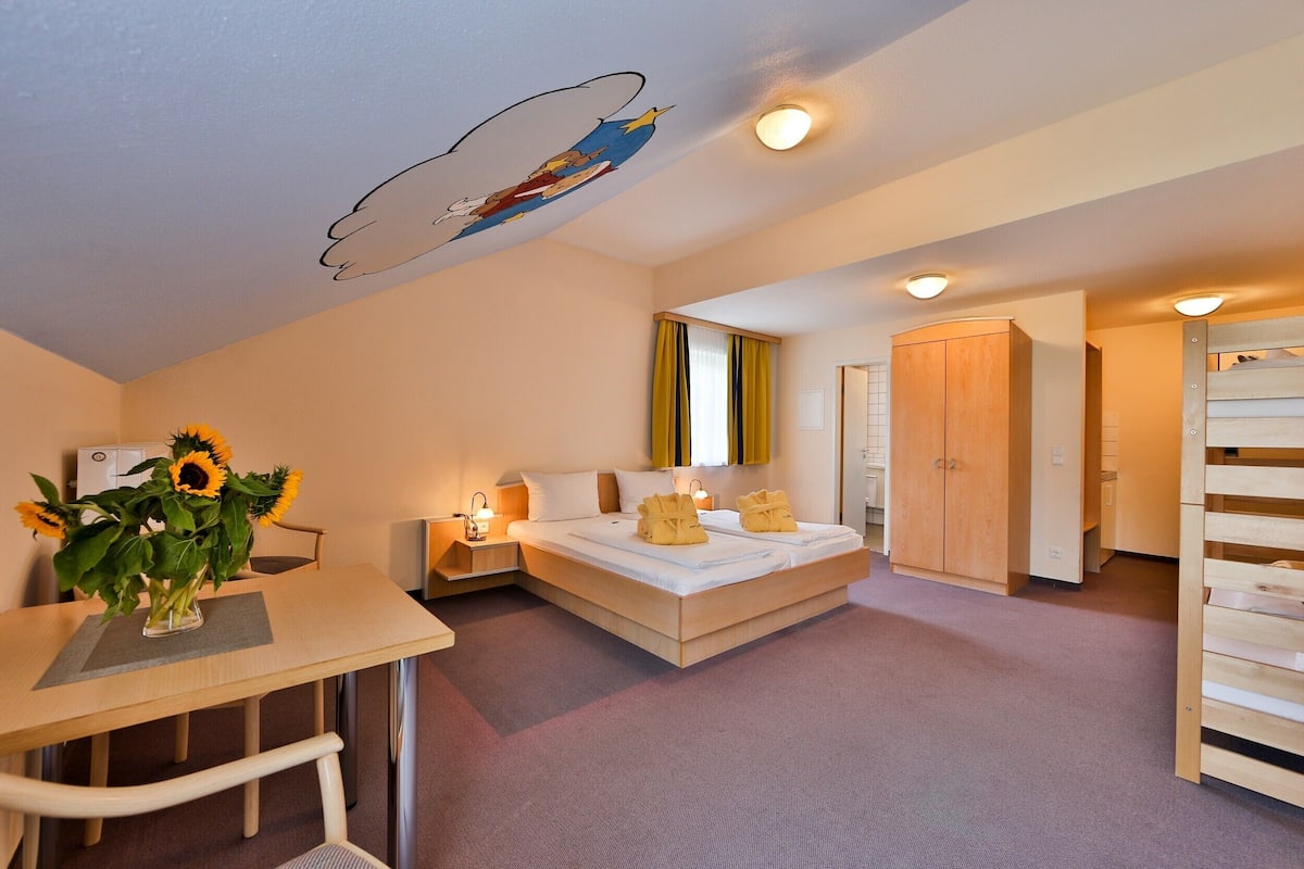 Ein Schlafzimmer mit einem Bett, Nachttischen, einem Schreibtisch mit einer Vase mit Sonnenblumen und einem Wandbild.