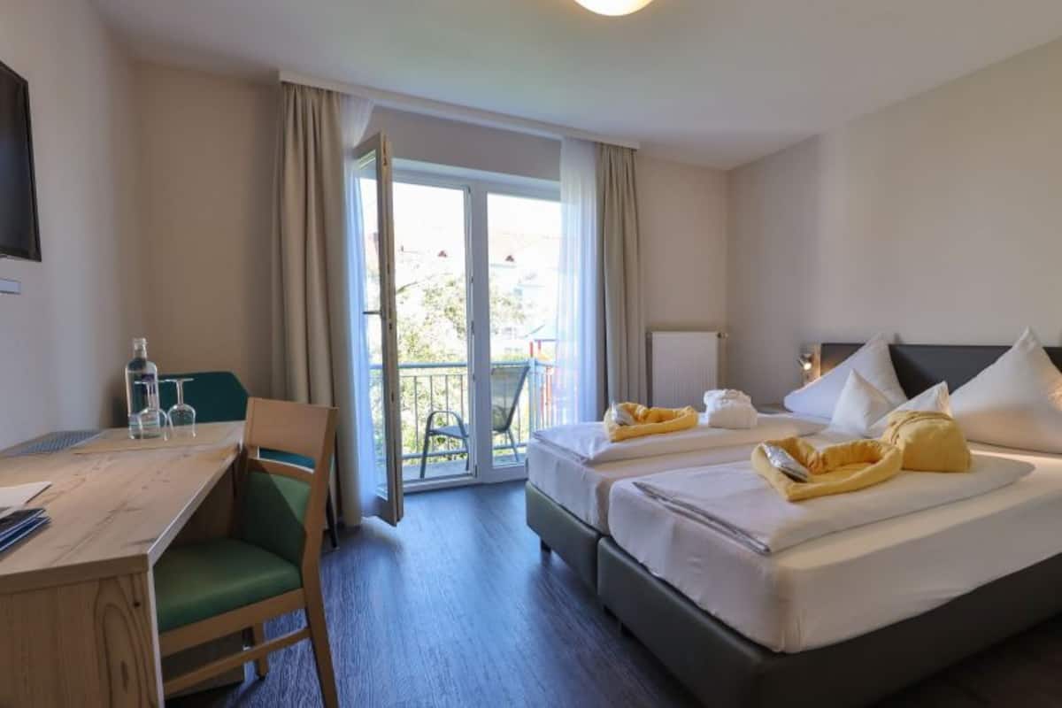 Ein Hotelzimmer mit zwei Betten, einem Schreibtisch, einem Stuhl, einem Balkon und Blick ins Freie.