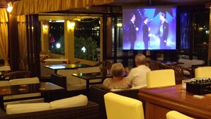 A bar area with wicker furniture and a television screen displaying a performance. - Maria' s Beach Apartments (Corfu)