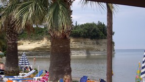 A beachside lounge area with lounge chairs, umbrellas, and a palm tree. - Maria' s Beach Apartments (Corfu)