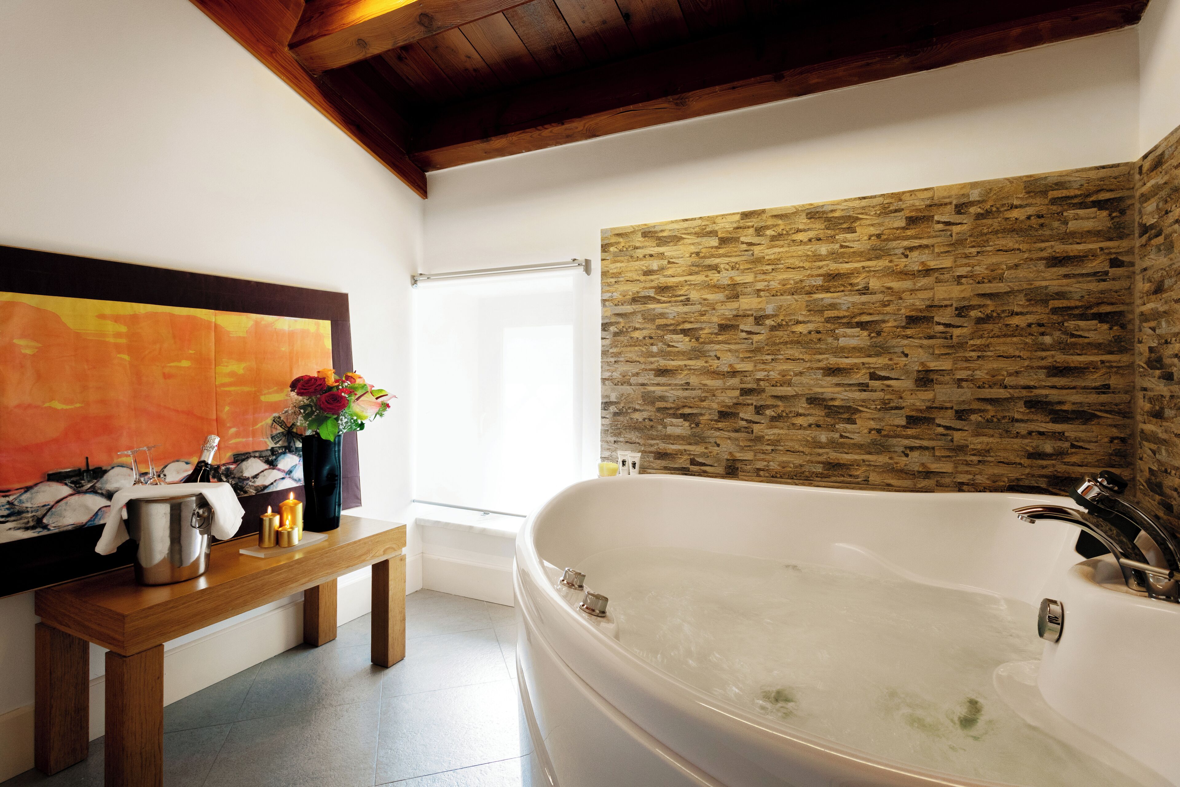 A modern bathroom with a freestanding bathtub, a wooden bench, and a stone wall.