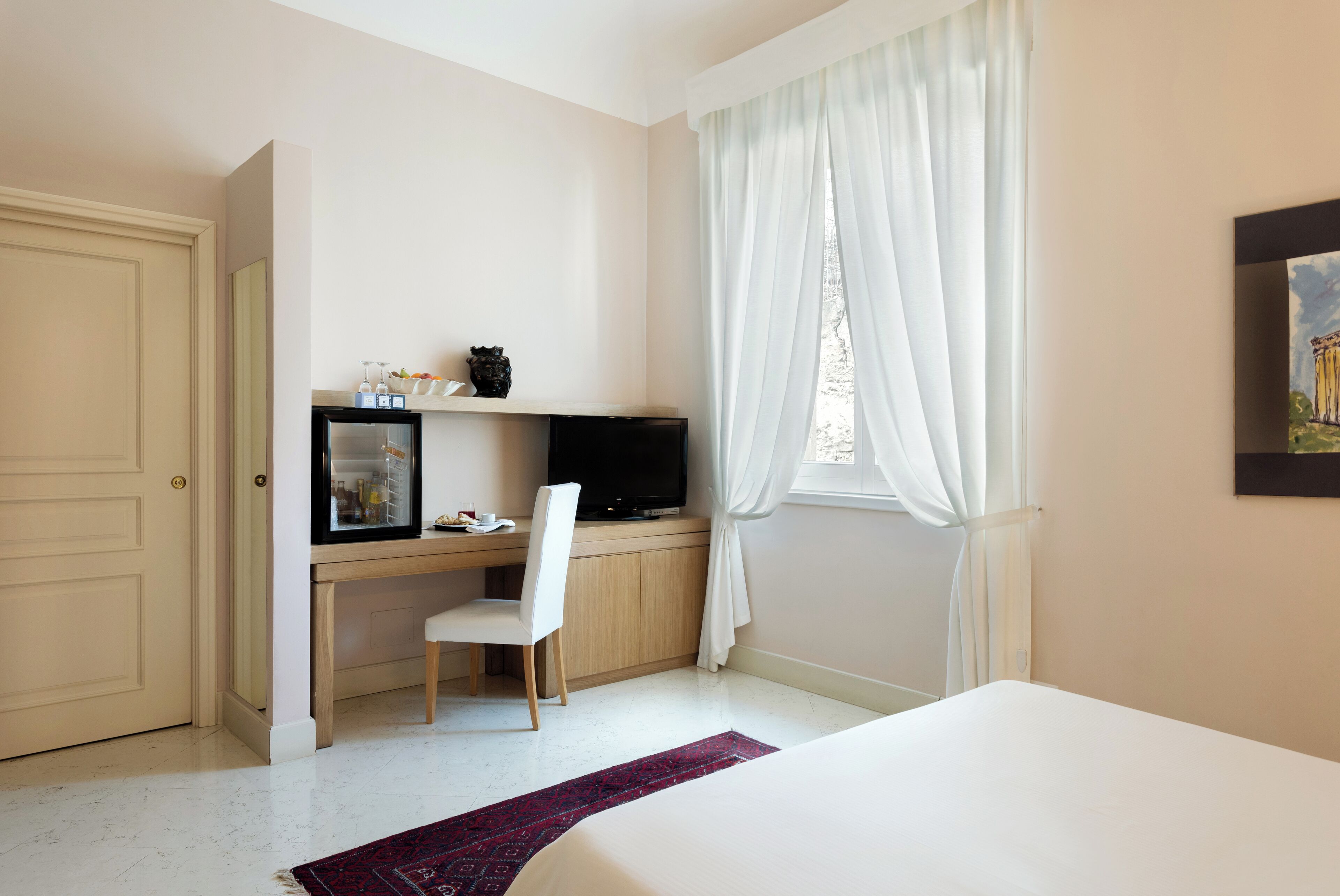 A hotel room with a bed, a desk with a computer, a chair, and a window with curtains.